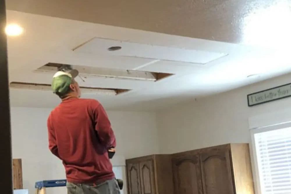 Man inspecting ceiling repair in a kitchen with exposed areas and wooden cabinets.